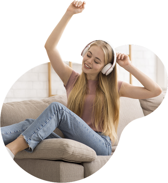 Jovem sentada confortavelmente em um sofá, usando fones de ouvido grandes e sorrindo com os olhos fechados, com os braços erguidos como se estivesse curtindo música.
